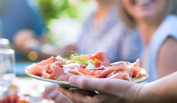 Close-up on a hand serving a dish of dry ham and cheese to friends gathered around a table in a garden to have fun together.