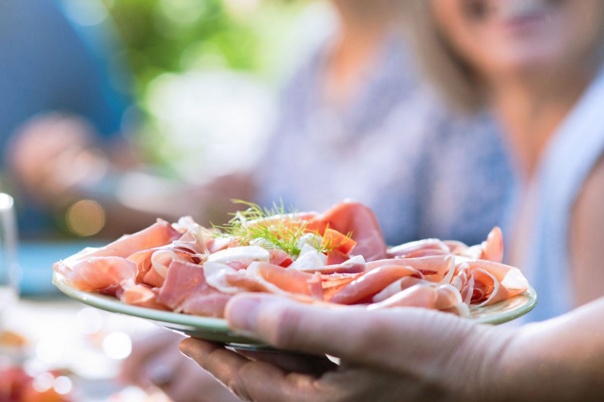 Close-up on a hand serving a dish of dry ham and cheese to friends gathered around a table in a garden to have fun together.